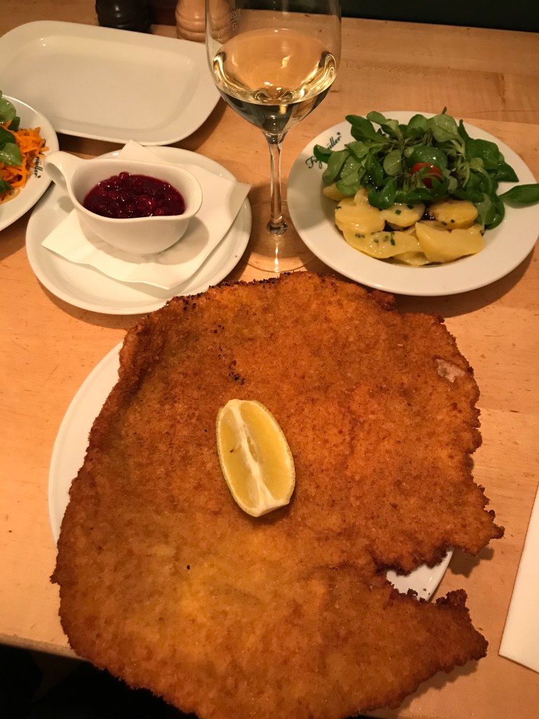 Large schnitzel with lemon on a plate next to a glass of white wine and potato salad