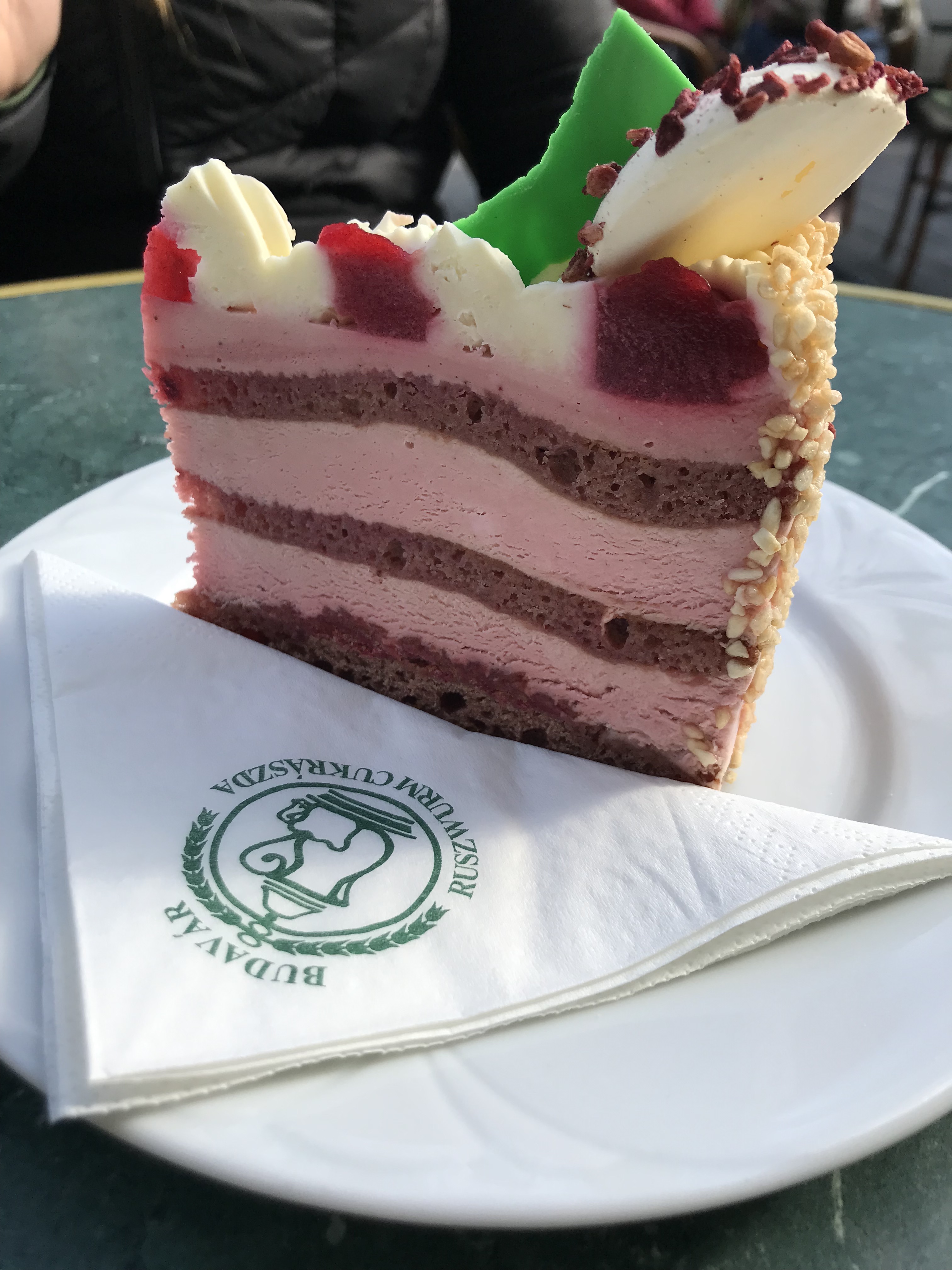 Cake slice with layers of pink and brown mousse. On top are white and red puffs of icing and a white macaron with red sprinkles.
