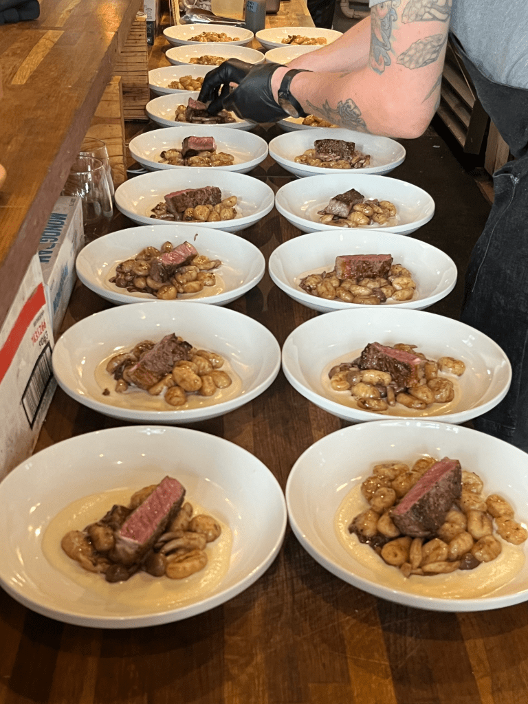 A dozen plates of gnocchi and steak lined up along a counter while a chef continues placing steak on top of the dishes